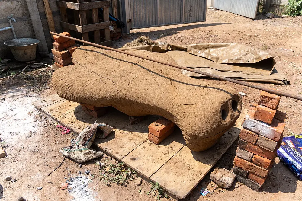 A long clay mould, cracked as it dries, lying on bricks in a workshop yard, with two round openings at one end.