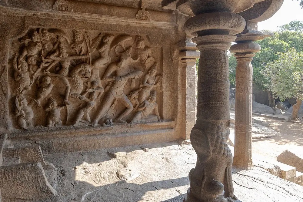A crowded granite relief inside a cave hall: on the left, a many-armed goddess rides a lion and leads an army; on the right, a giant warrior with a buffalo's head draws back with a club. A carved pillar with a seated lion at its base stands in front.