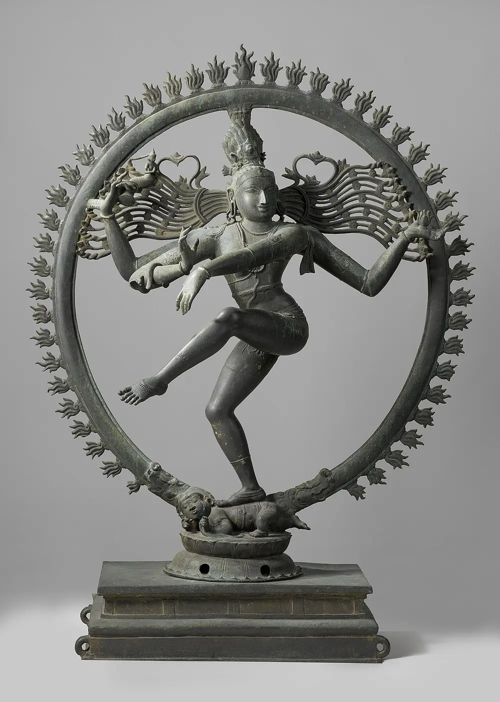 A dark metal statue of four-armed Shiva dancing on one leg inside a large ring edged with flames. His hair flies out sideways, one foot rests on a small crouching figure, and the other leg is lifted across his body.