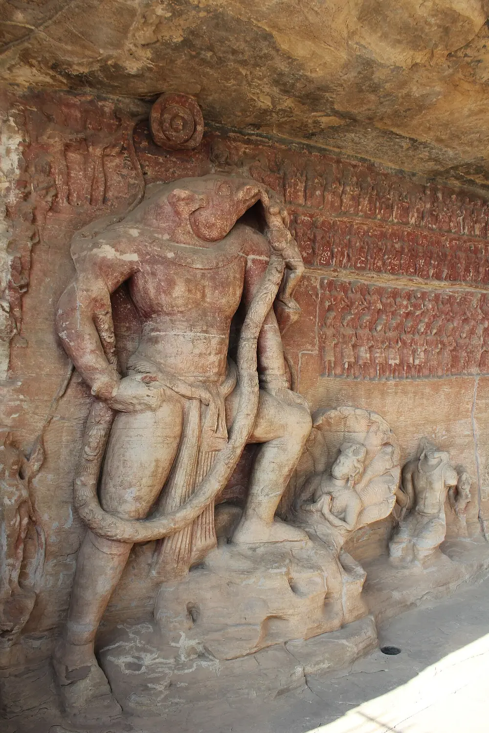 A huge relief in a rock face of a figure with a man's body and a boar's head, one foot raised on a rock, with a small female figure hanging from his tusk and rows of tiny carved figures filling the wall behind him.