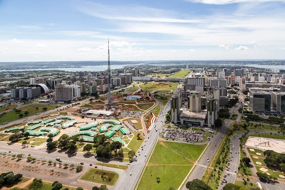 An aerial view of a modern city with a very wide central avenue of lawns, government buildings in a row and a tall TV tower, with a lake in the distance.