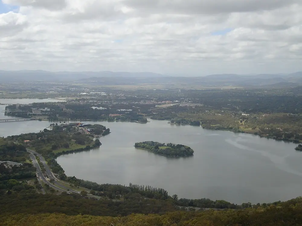 An aerial view of a large lake with an island, surrounded by low green city suburbs and wooded hills.