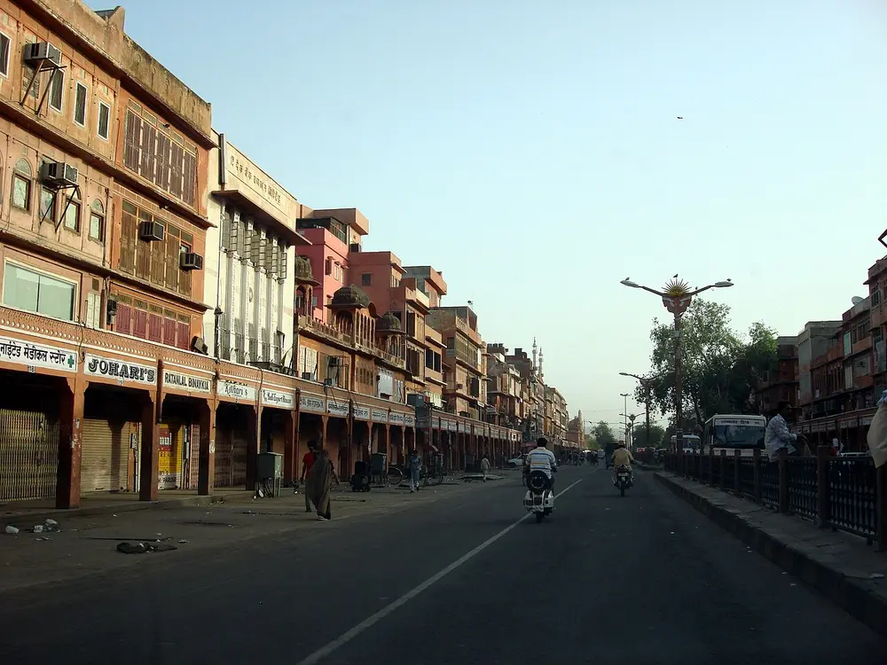 A wide, straight road in old Jaipur lined on both sides by matching pink-red buildings with shops below and balconies above.