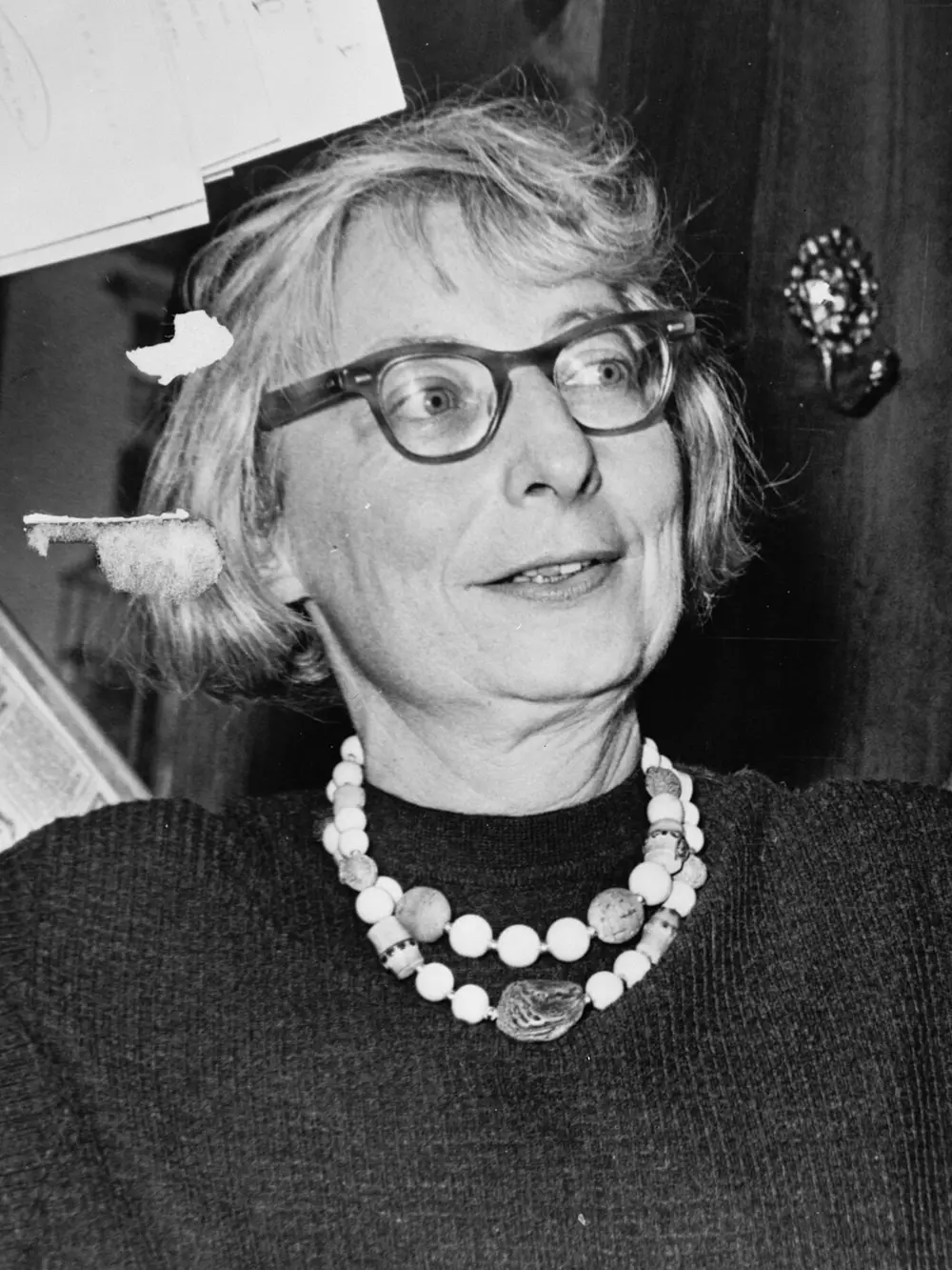 A black and white photo of a woman with short grey hair, large round glasses and a chunky bead necklace, at a press conference.