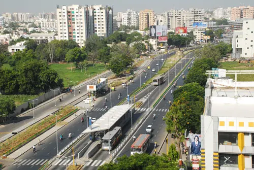A wide city road seen from above, with a glass-roofed bus station and red buses in a separate lane in the middle of the road.