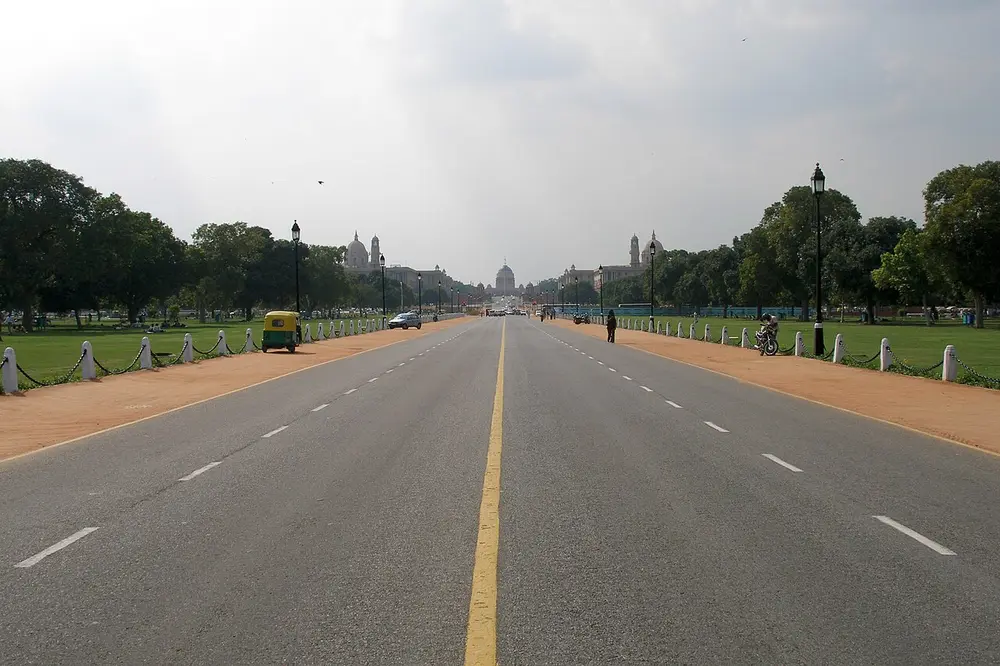 A long, straight, wide road with lawns and trees on both sides, leading to large domed government buildings in the far distance.