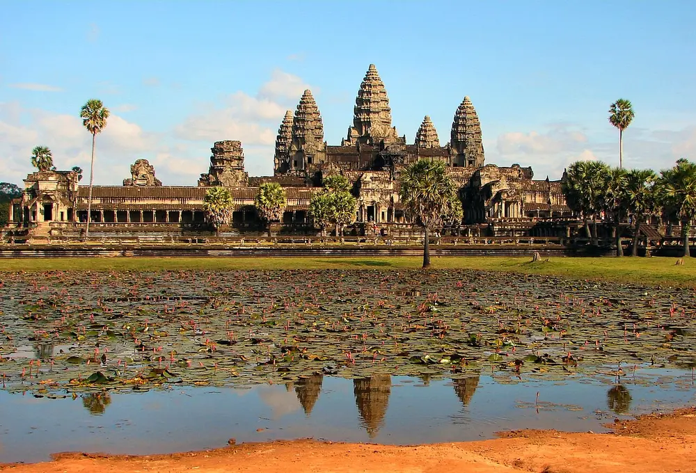Angkor Wat, a grey stone temple with five tall towers shaped like lotus buds, behind long galleries and palm trees, reflected in a pond with water lilies in front.