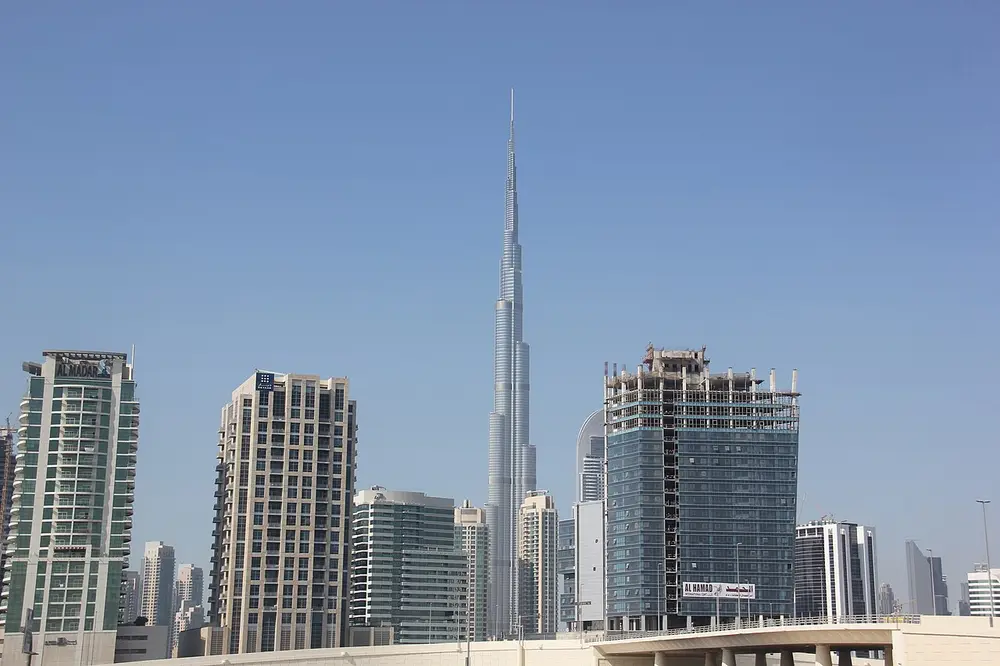 The Burj Khalifa in Dubai, a very tall, slim silver tower stepping back as it rises to a needle-like spire, more than three times as tall as the tower blocks around it, under a clear blue sky.