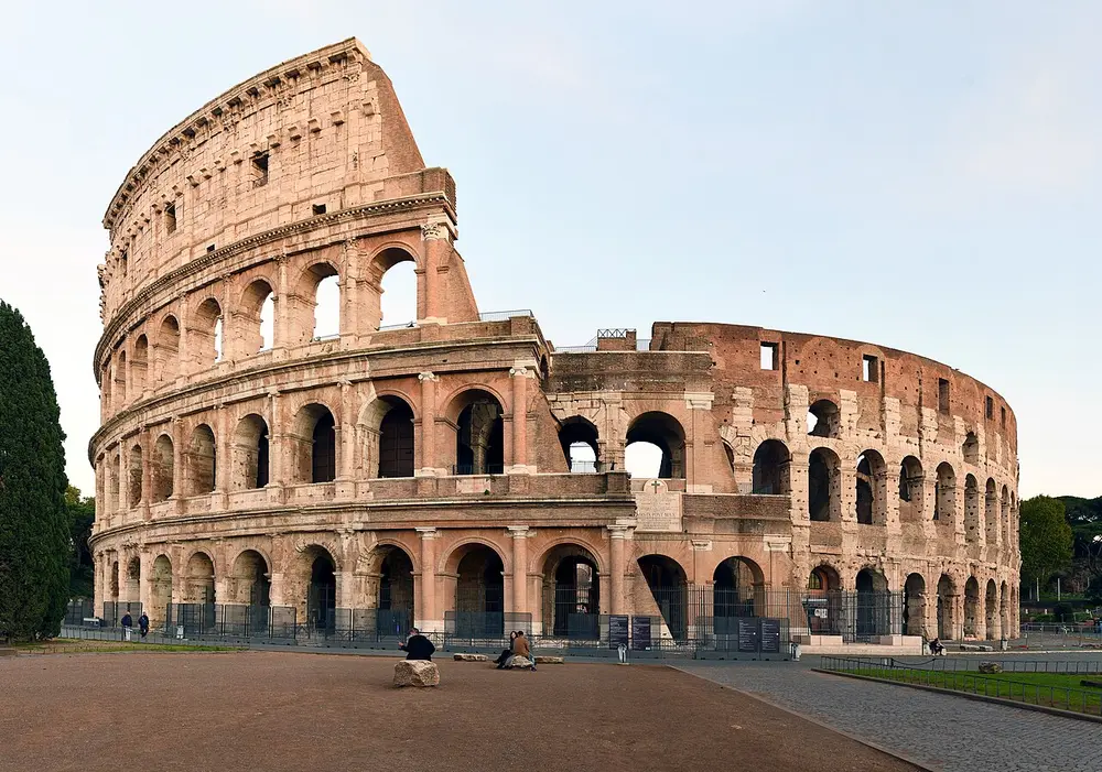 The Colosseum in Rome, a huge oval stone arena with three rows of arches and a solid top storey, part of its outer wall broken away to show inner rings of arches.