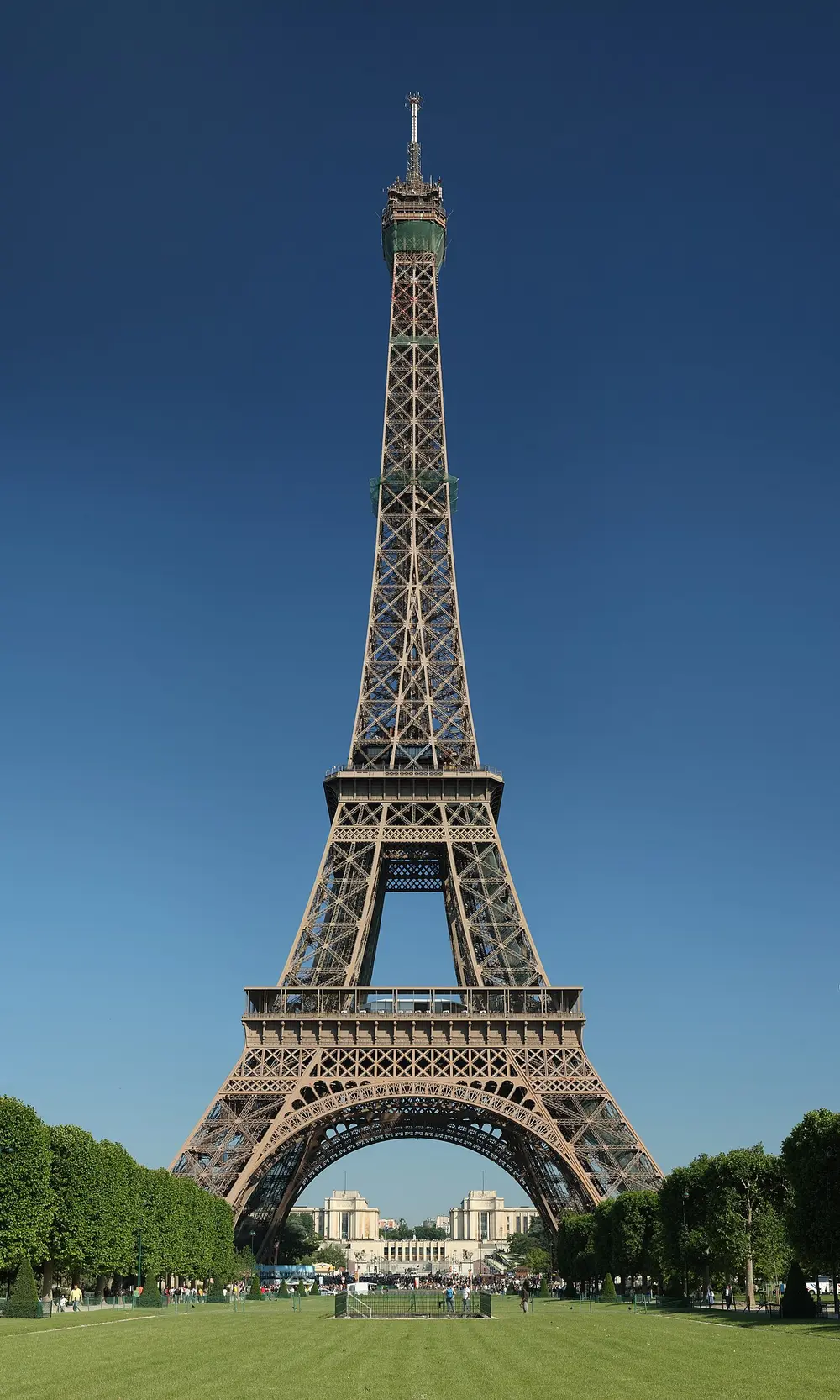 The Eiffel Tower in daylight against a deep blue sky, a tall tapering lattice of brown iron with a wide arch between its four legs, standing on a long green lawn.