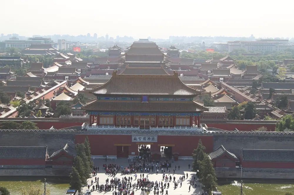 The Forbidden City in Beijing seen from a hill to the north: a two-roofed gate tower above a long red wall and a moat, with crowds at the gate and rows of palace halls with golden-yellow tiled roofs stretching away behind.