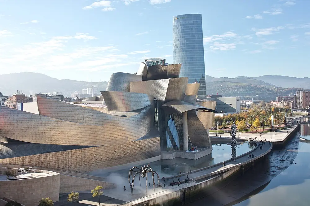 The Guggenheim Museum in Bilbao, a cluster of swooping, curved silver metal forms and glass beside a river, with a giant spider sculpture on the riverside walkway and a glass tower behind.
