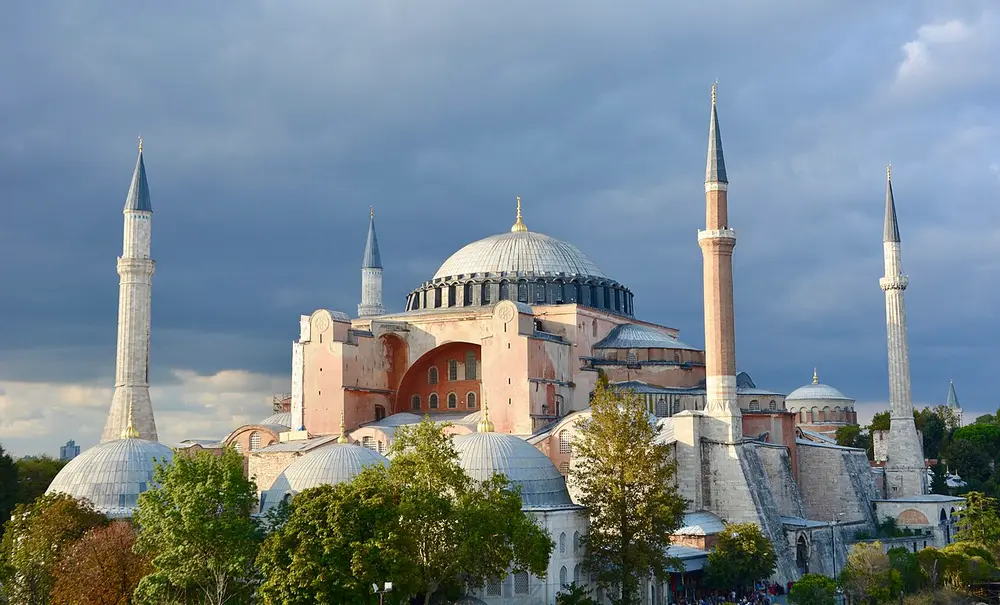 Hagia Sophia in Istanbul, a massive pink and cream building with a huge, shallow central dome ringed with small windows, smaller domes and half-domes around it, and four tall thin minarets.