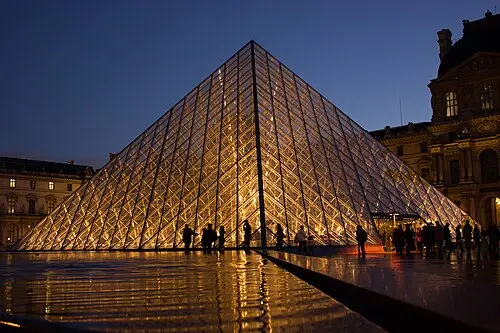 The Louvre Pyramid at dusk, a tall pyramid of glass panes held in a metal grid, glowing from inside, with the old stone palace of the Louvre behind and people walking past.