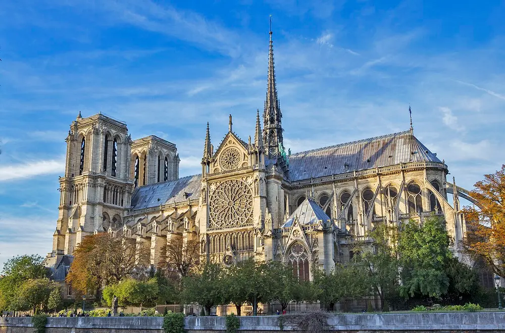 Notre-Dame cathedral in Paris in autumn sunlight, with two square towers on the left, a huge round rose window on its side, a tall thin spire, and stone arms of flying buttresses along the right.