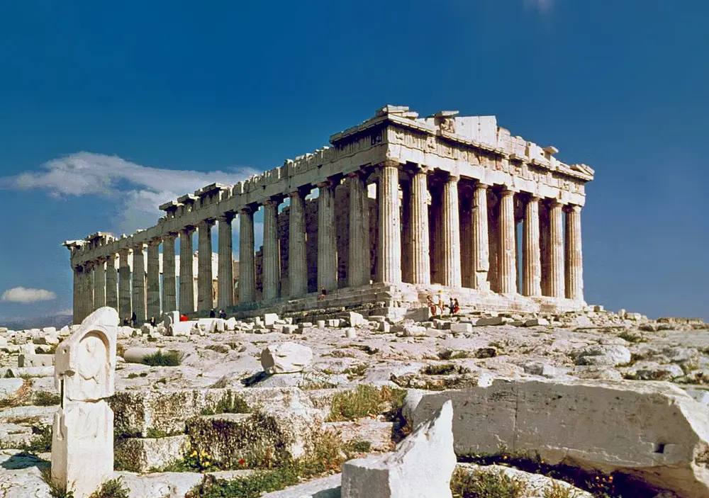 The ruined marble Parthenon on a rocky hill, a long rectangular temple surrounded by rows of thick fluted columns, with part of its roof and triangular end missing, under a deep blue sky.