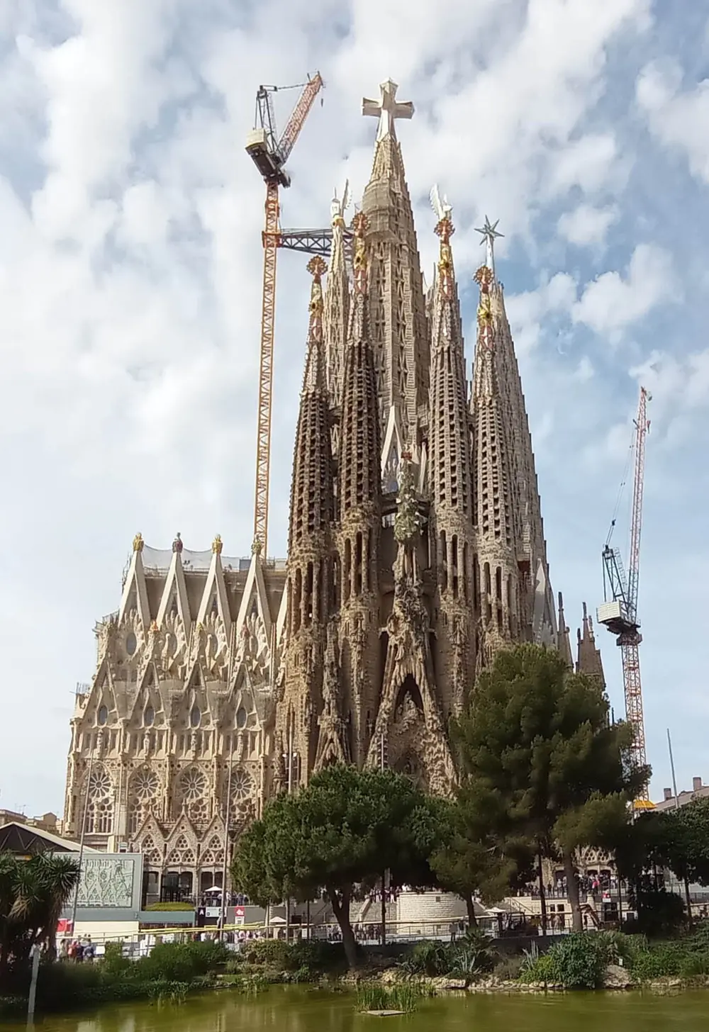 The Sagrada Família basilica in Barcelona, a group of very tall, thin, honeycombed stone spires topped with crosses and coloured finials, with tall cranes beside it.