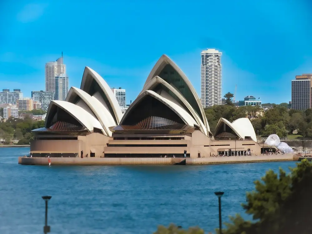 The Sydney Opera House on the harbour, a group of white curved shell roofs like sails or segments of an orange, on a broad base, with city towers behind and blue water in front.