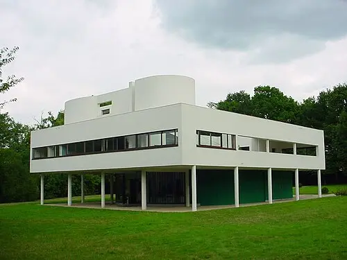 Villa Savoye, a white box-shaped house raised on thin round columns above a green lawn, with a long strip of windows across its upper floor and curved white walls on the roof.