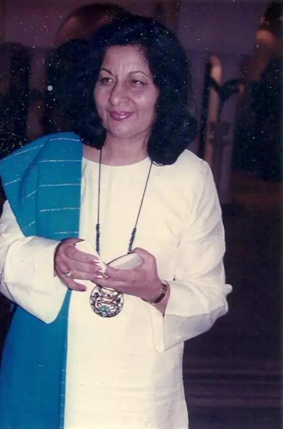 A photograph of Bhanu Athaiya with dark curly hair, in a white outfit with a blue scarf and a large round pendant, holding a small object in her hands.