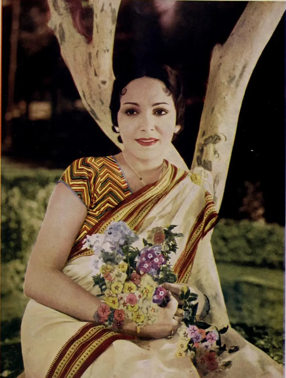 A hand-coloured 1938 publicity photograph of Devika Rani in a cream sari, seated against a tree trunk and holding a bunch of flowers.