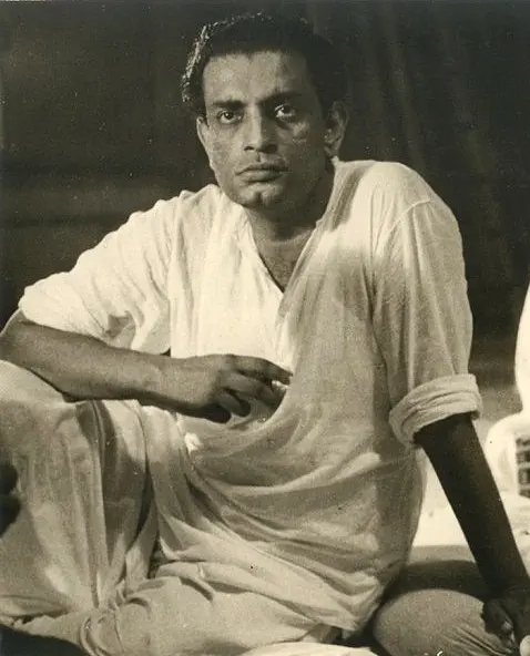 A black and white photograph of a young Satyajit Ray in a white kurta, sitting on the floor with one arm resting on his knee, during the recording of music for Pather Panchali.