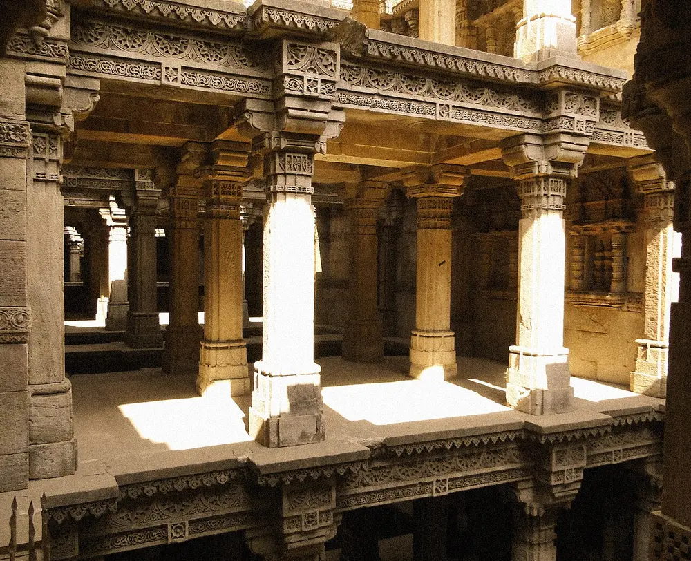 Inside the Adalaj stepwell: carved stone pillars, brackets and beams frame open galleries on several levels, with sunlight falling on the stone floors.