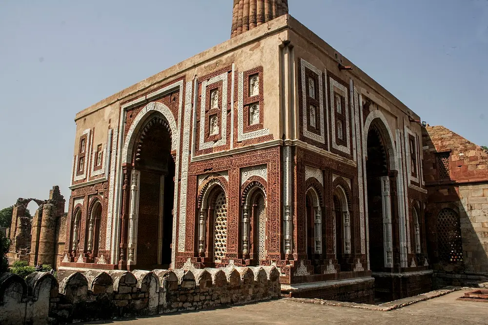 A square red sandstone gatehouse with a tall pointed arch on each face, its walls trimmed with white marble bands and lattice windows, with the Qutb Minar rising behind.