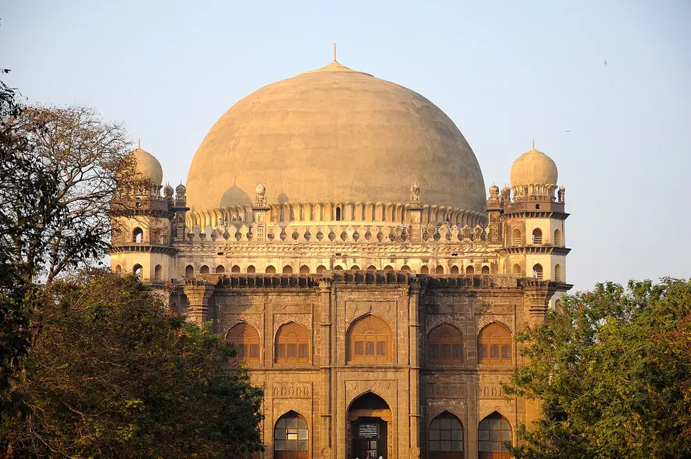 The Gol Gumbaz, a massive cube-shaped tomb with a gigantic round dome on top and a domed seven-storey tower at each corner, lit by evening sun.