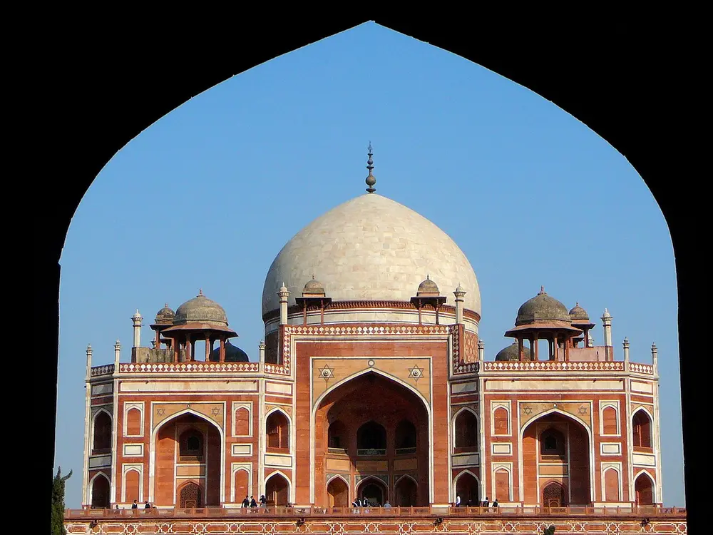Humayun's Tomb, a red sandstone building with white marble borders, tall arched portals and a white marble dome flanked by small domed kiosks, framed by the pointed arch of a gateway.