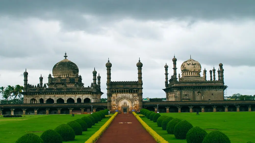 Two richly decorated buildings facing each other across a garden, each with a bulbous dome and many slender minarets, at the end of a long red path lined with clipped hedges.