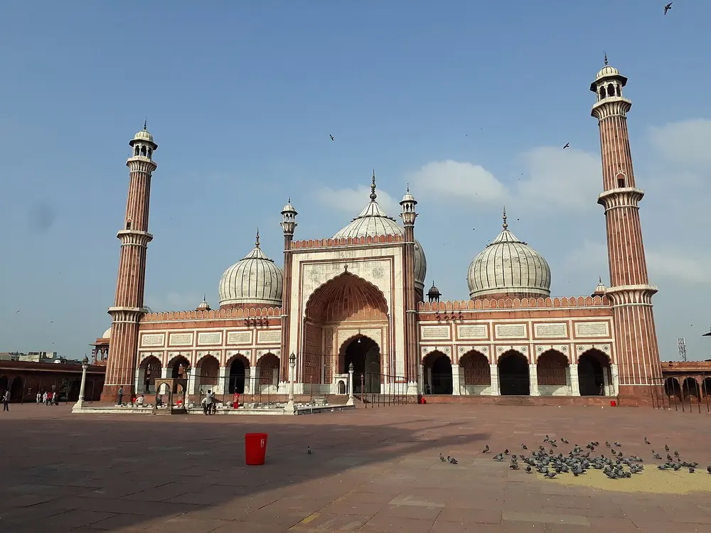 The Jama Masjid, a red sandstone mosque with a tall arched central gateway, three white marble domes with thin dark stripes, and two tall striped minarets, behind a big paved courtyard.