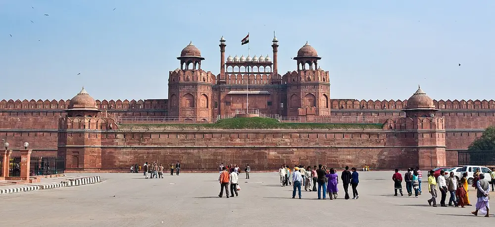 The long red sandstone walls of the Red Fort with the Lahori Gate in the centre, topped by small white domed kiosks, seen across a wide open square.