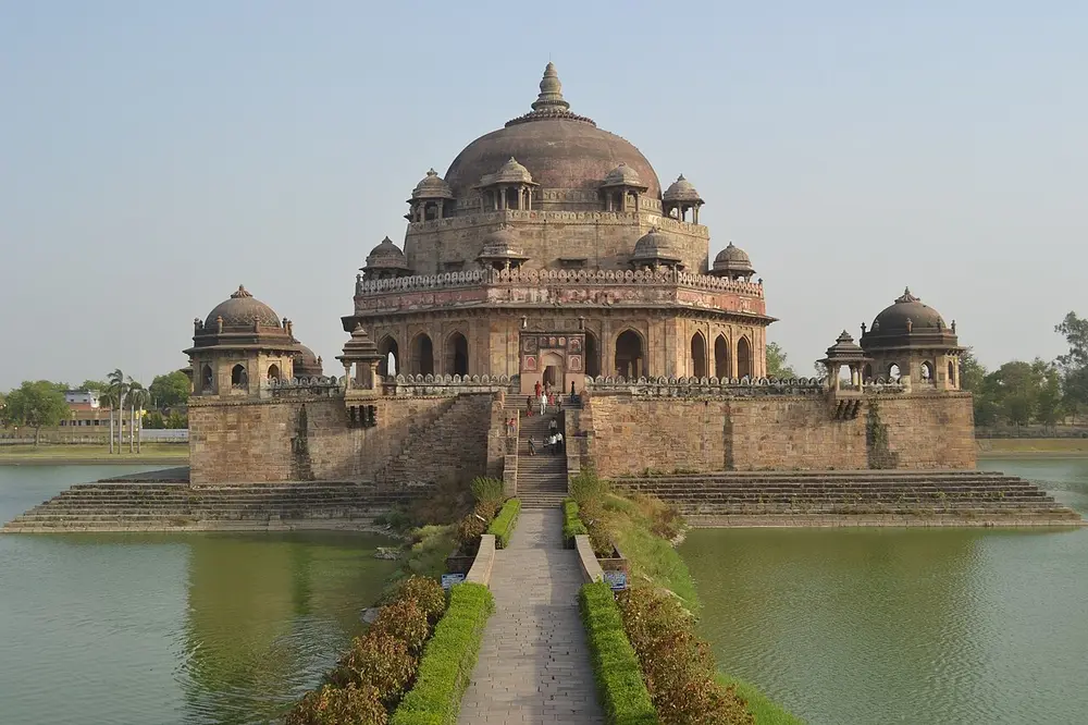 A large octagonal sandstone tomb with a big dome and many small domed kiosks, standing on a square platform in the middle of a lake, reached by a long path.