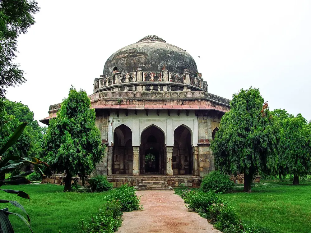 An octagonal stone tomb with a row of pointed arches around its base, a ring of small turrets and a large grey dome, among trees in a garden.