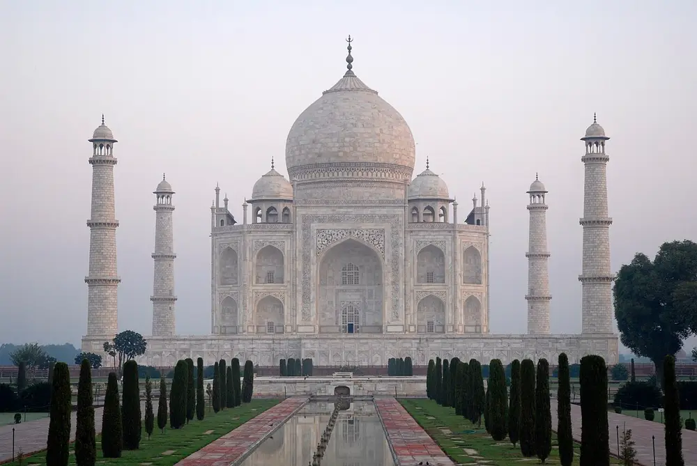 The white marble Taj Mahal in soft morning light, with its large onion-shaped dome, smaller domes, and four slim minarets at the corners, seen down a long garden pool lined with cypress trees.