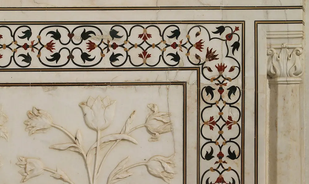Close-up of a white marble wall at the Taj Mahal: a border of tiny red, green and black stone flowers set flat into the marble, beside a carved marble relief of tulip-like flowers.