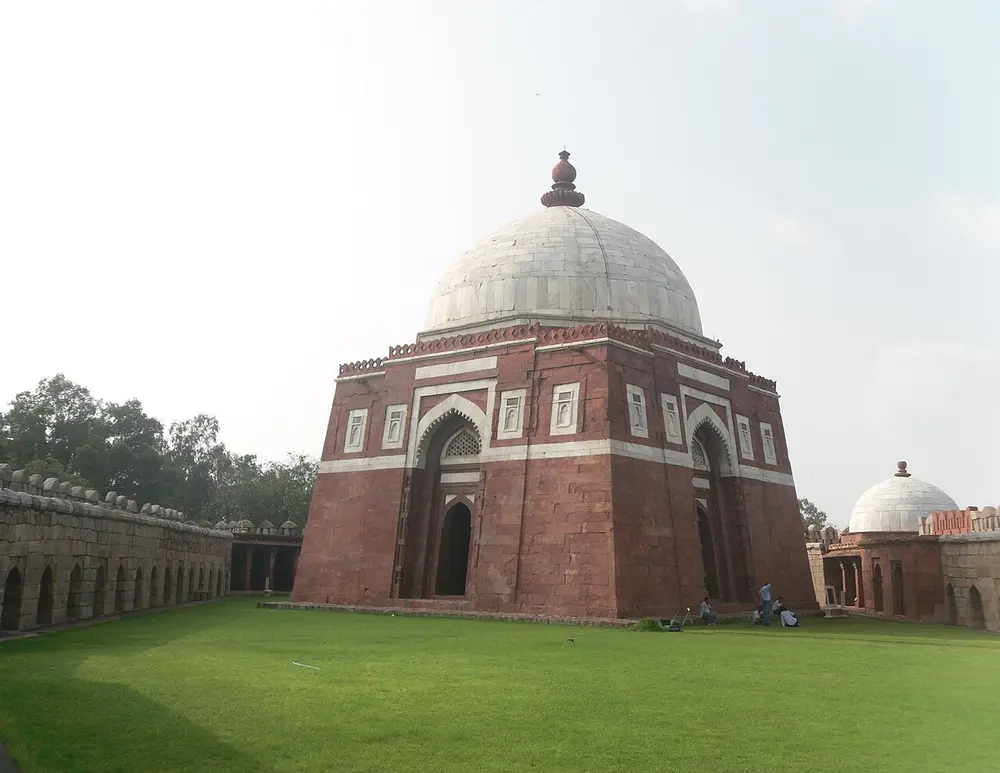 A square tomb of red sandstone with walls that slope inward, white marble bands around its arches and a white marble dome, standing in a walled lawn.
