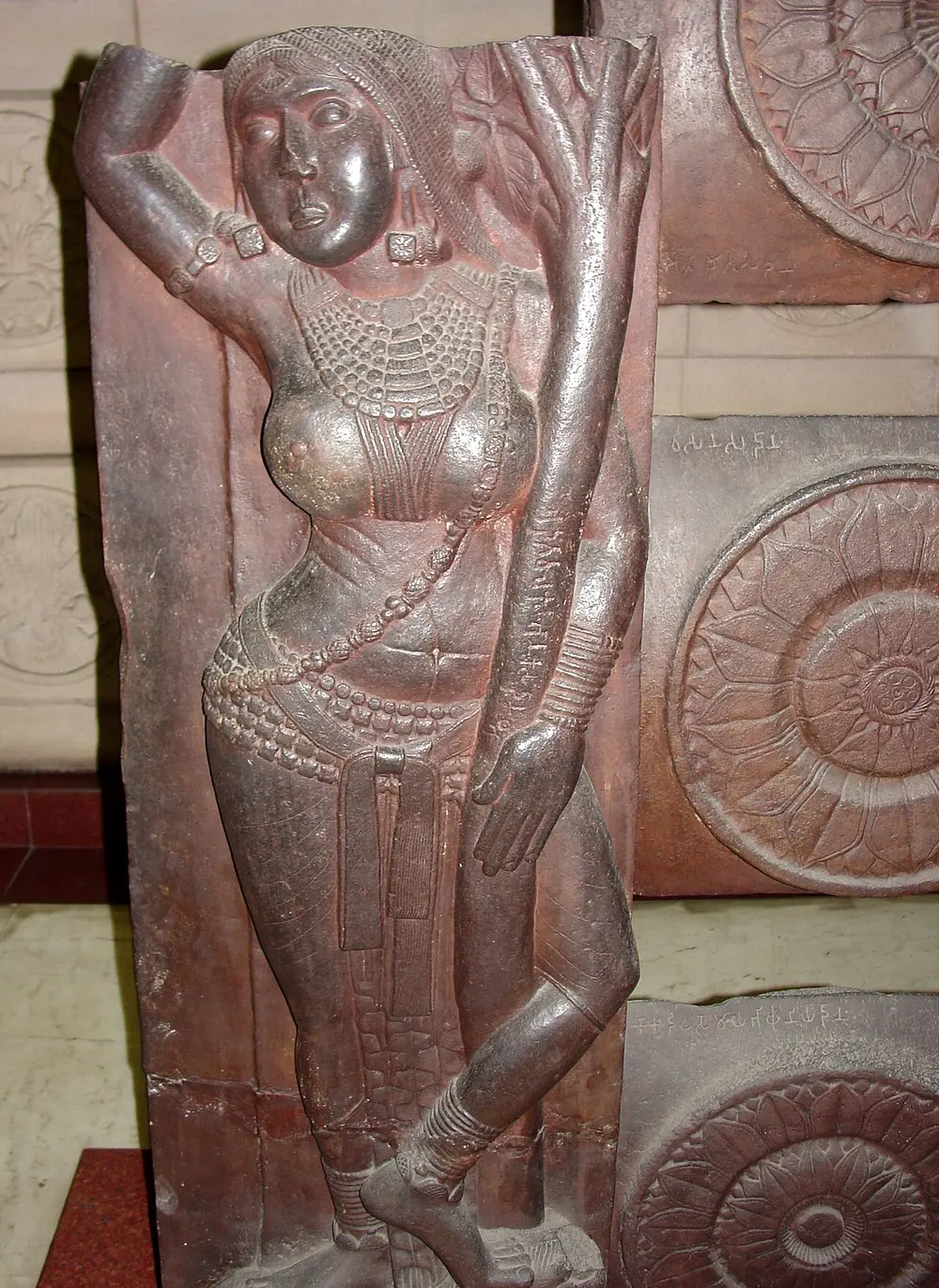 A tall red sandstone railing post carved in low relief with a woman in heavy jewellery standing under a tree, holding a branch above her head and wrapping one leg round the trunk.
