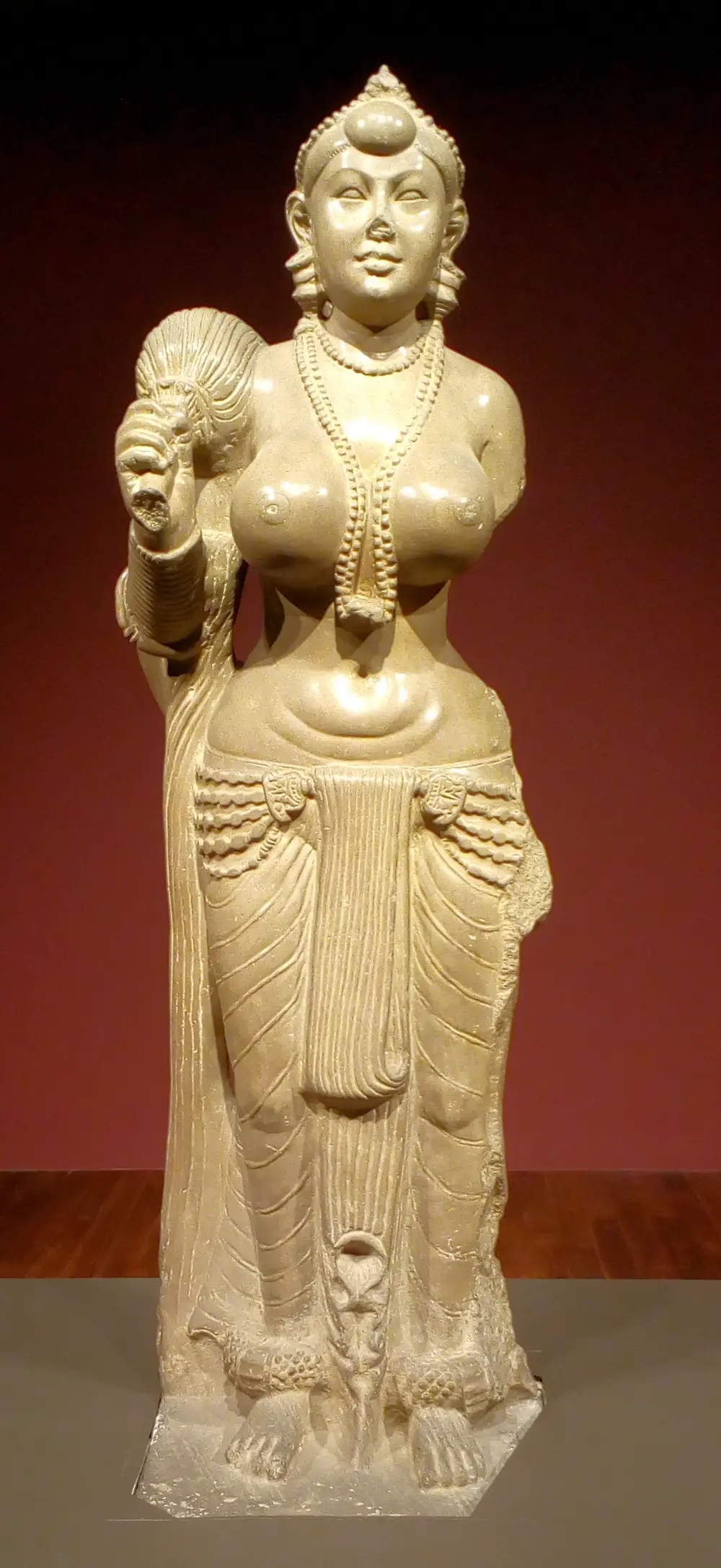 A life-size, highly polished cream stone statue of a standing woman holding a fly-whisk over her right shoulder, wearing heavy necklaces, a beaded belt and a finely pleated lower garment, against a dark red wall.