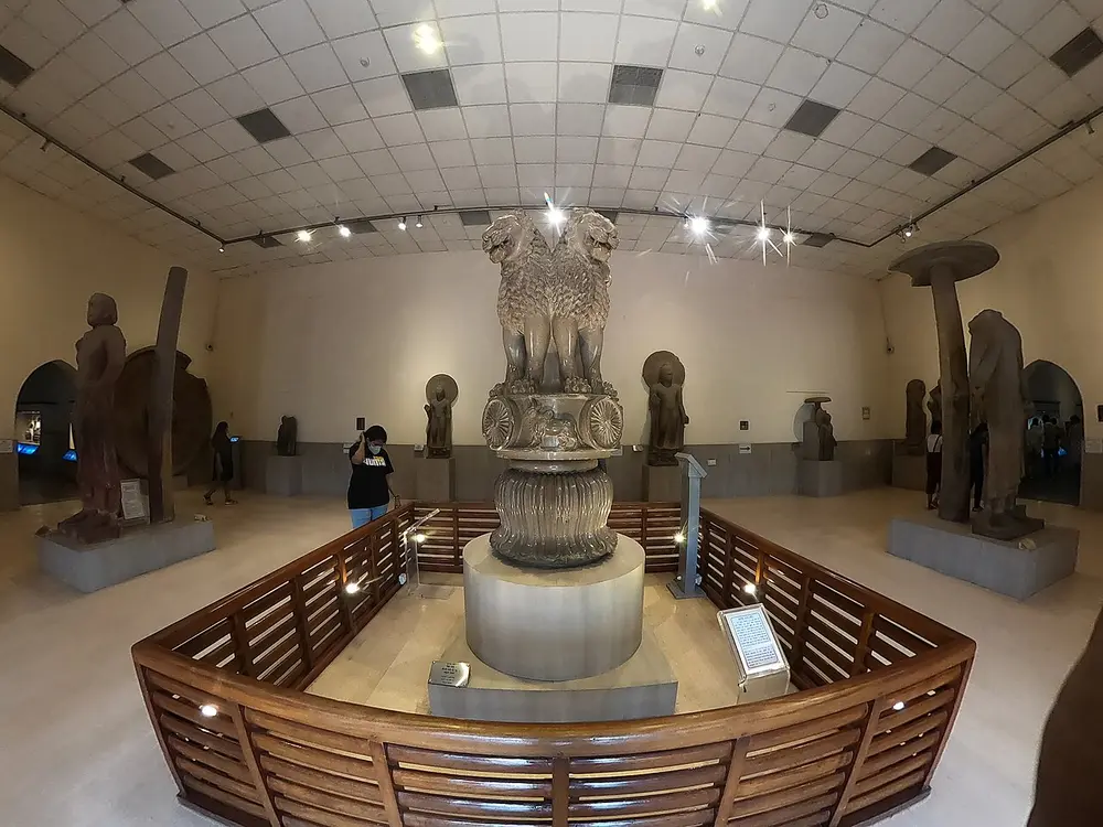 A museum hall with the Sarnath Lion Capital in the centre on a round stand behind a wooden railing: four lions back to back on a carved drum above a bell-shaped lotus, with other ancient sculptures around the walls.
