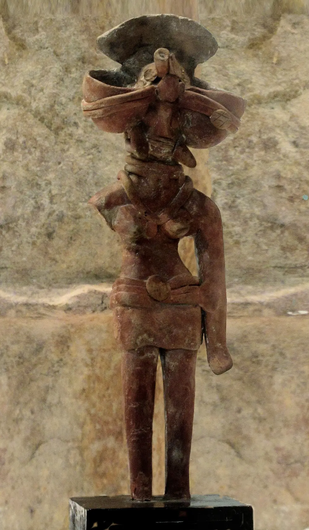 A standing clay figurine of a woman wearing a large fan-shaped headdress, heavy necklaces and a short skirt with a wide belt.