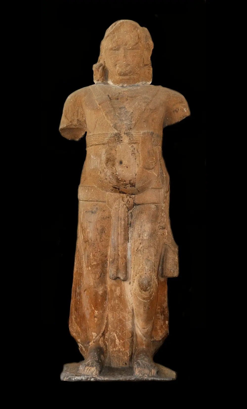 A tall, heavy stone figure of a standing man with broken arms and a damaged face, wearing a scarf and a long lower garment, against a black background.