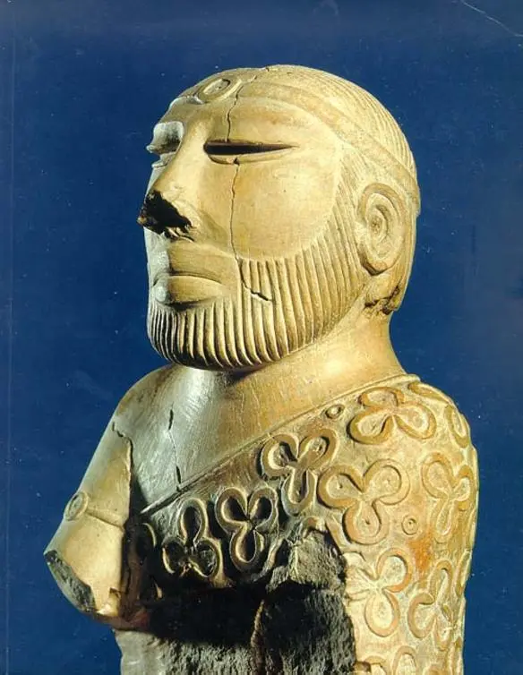 A pale stone bust of a bearded man with half-closed eyes and a band round his head, wearing a robe over one shoulder decorated with raised trefoil patterns.