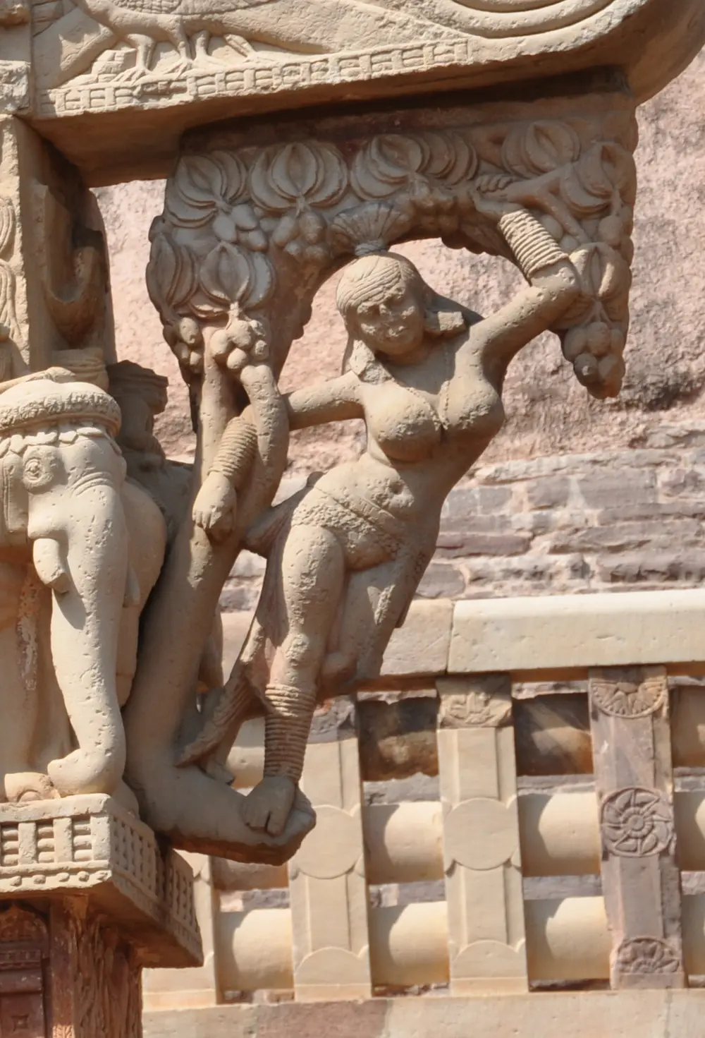 A carved stone yakshi swinging out from a gateway, holding a tree branch above her with one arm, her body curving away from the post beside carved elephants.