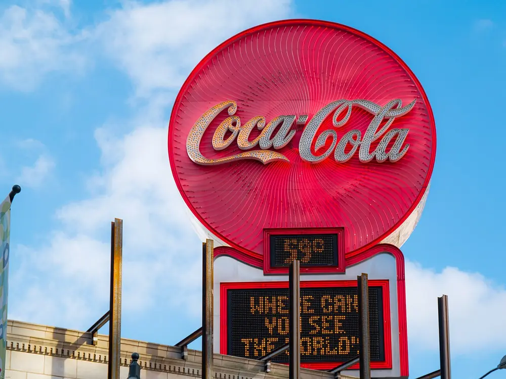 A large round red neon sign with the words Coca-Cola in flowing white joined-up handwriting, on a rooftop against a blue sky, above a small electronic board.