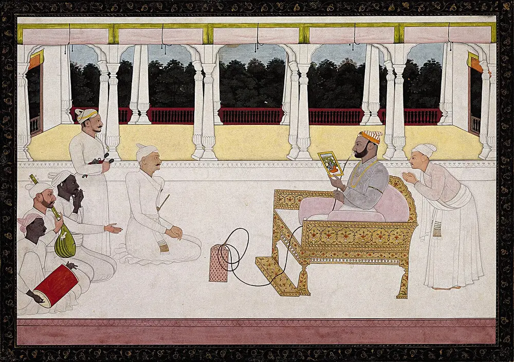 A turbaned ruler sits on a low golden seat on a white terrace, studying a small painting, while attendants and musicians sit or stand around him.