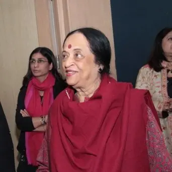 Anjolie Ela Menon, an older woman with dark hair tied back and a red bindi, wearing a red shawl, smiling at an event.