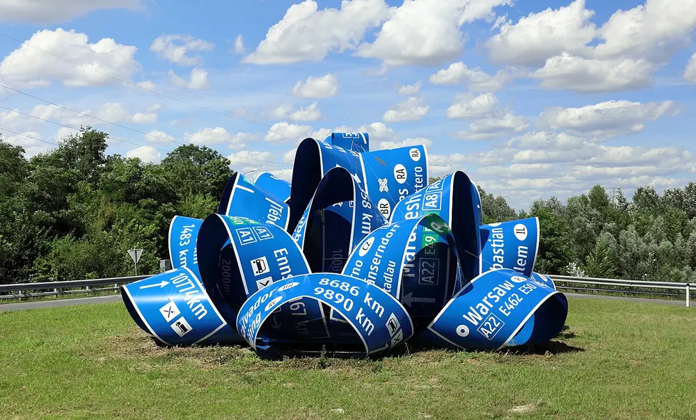 A large outdoor sculpture on grass beside a road, made of blue motorway signs curled into loops, with place names and distances such as Warsaw and Mumbai.