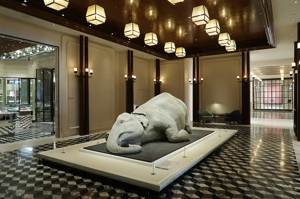 A life-size grey elephant lying down with its head on the floor, covered all over in small bindis, displayed in a hotel lobby.